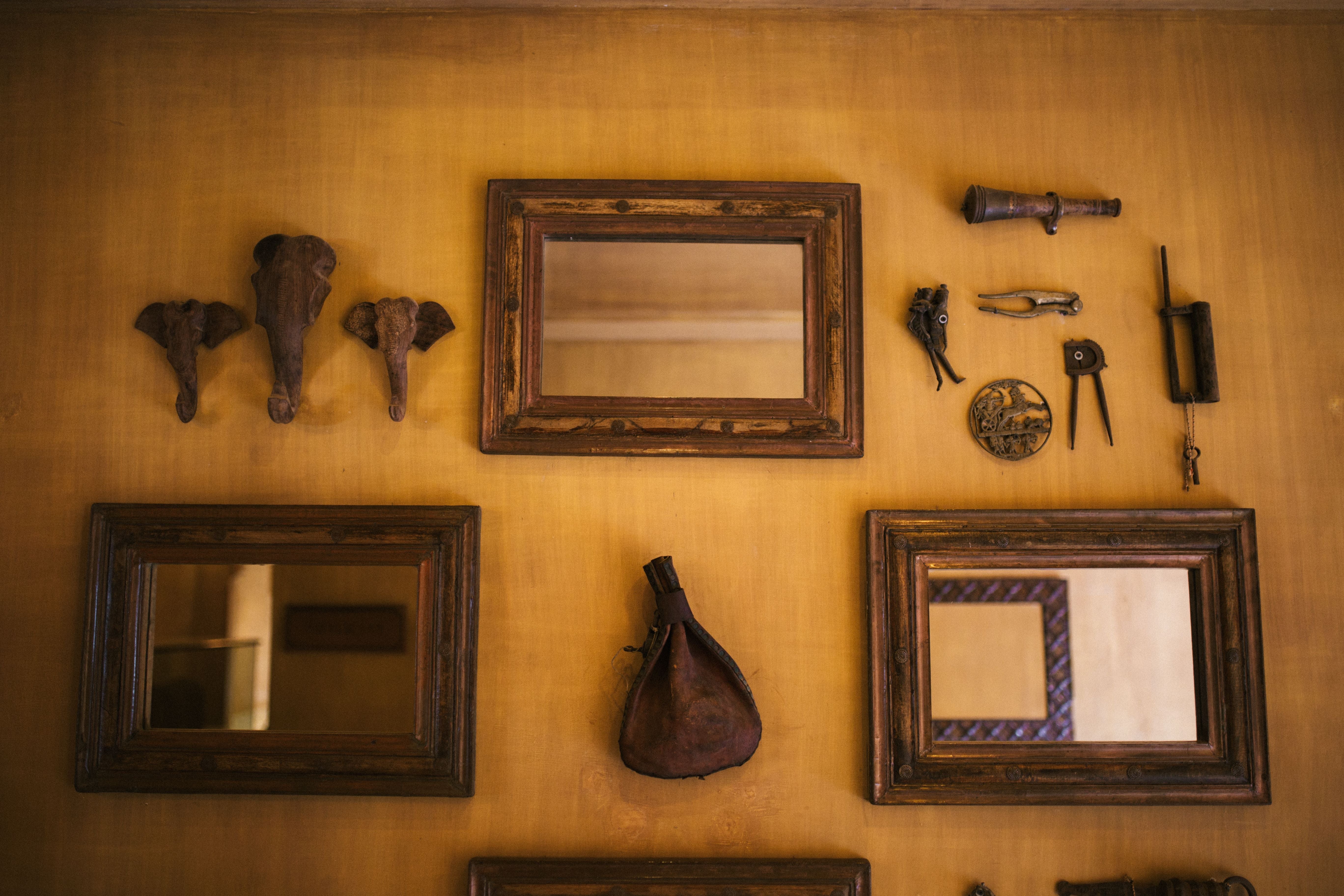 A warm, rustic wall display featuring multiple vintage dark wood picture frames, elephant head wall mounts, a small hanging leather bag, and antique metal tools against a textured ochre-yellow background.